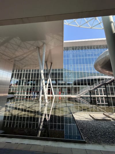 The image shows the Rho Fiera Milano complex in Rho, Italy. The view is looking up at the architecture from an interior perspective. A geometric glass and metal structure is visible, along with part of a large, modern building featuring a reflective facade of glass windows. A pool of water is in the frame, reflecting the building's exterior and the sky. Metal support beams are prominent in the foreground. The sky is clear and blue.