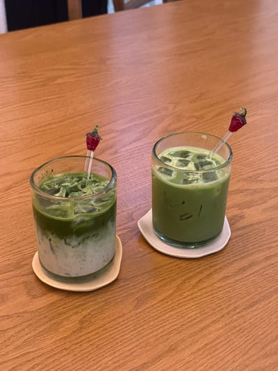The image displays two iced green beverages on a light-colored wooden table. Each drink is contained within a clear glass tumbler and rests on an individual, irregularly shaped, light beige coaster. The beverage on the left exhibits a distinct two-layered separation: a milky white base liquid topped with a darker green liquid and ice cubes. The beverage on the right is a uniform light green color with ice cubes throughout. Both glasses contain a clear glass stirrer, each topped with a small, red, chili-shaped decoration. The wooden table shows horizontal grain patterns. The background is blurred, indicating an indoor setting consistent with a cafe or restaurant, but provides no specific identifying features. No individuals are visible. The precise location within the city of Guadalajara, Mexico, cannot be determined from the provided image.