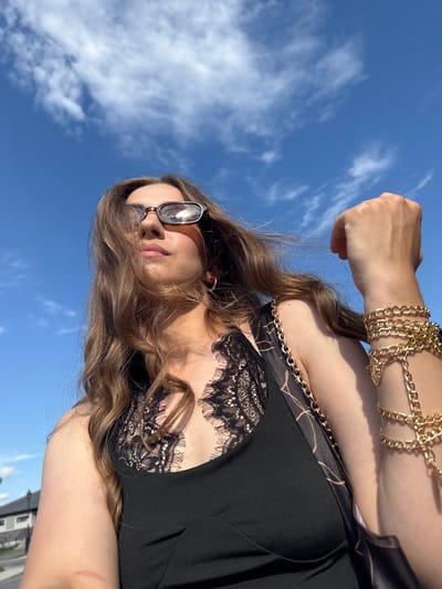 A young woman wearing sunglasses, a black lace-trimmed top, and a gold chain bracelet is captured in a low-angle, close-up shot outdoors. Her long, wavy brown hair is windswept, and she is looking upwards against a bright blue sky with scattered white clouds. The background shows a glimpse of suburban houses and a street, suggesting a location in Gatineau, Canada. The overall lighting indicates it is daytime, possibly late morning or afternoon, given the bright sunlight and shadows. The woman's left arm is bent, with her fist clenched near her head, and her right arm is out of frame but adorned with the prominent gold bracelet.