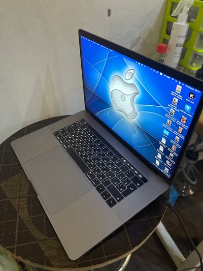 An angled, medium shot captures a silver Apple MacBook Pro laptop placed on a dark, circular table with a patterned surface. The laptop is open, displaying a desktop with the Apple logo as the wallpaper and various icons. The keyboard is visible, featuring Cyrillic characters. To the right of the laptop, a spray bottle labeled "simple" and "HOME" is on a green shelf unit. The background shows a textured, off-white wall with shadows. The scene appears to be an indoor setting, potentially a home or office. The city of Oskemen, Kazakhstan is not depicted or implied in the image.