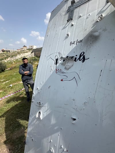 The image captures a scene in Dura, Palestinian Territory, centered on a damaged, light-colored metal panel positioned outdoors. The panel displays numerous indentations, perforations, and torn metallic edges, consistent with projectile or impact damage. A piece of crumpled metallic debris is affixed to its surface. A black line art illustration on the panel depicts one hand applying red nail polish to the nails of another hand. Above this artwork, the cursive text "But first" is legible, with the word "nails" partially obscured below. A metal fixture is attached to the panel's upper section. To the left of the panel, a bearded male subject stands with his arms crossed, wearing a dark patterned sweater and dark trousers, looking towards the camera. He is situated on uneven ground covered with grass and sparse vegetation, where a red-and-white striped barrier tape is visible in the foreground. The background features a sloping landscape with scattered rocks and several residential buildings, one of which includes a dark, domed architectural feature. The sky is clear blue with a few white clouds, and the scene is brightly lit by natural sunlight.