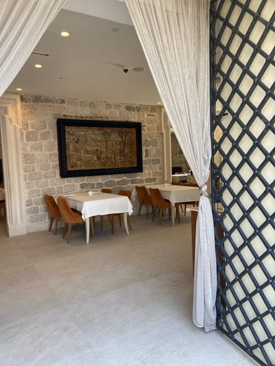 The image shows a view from an entryway looking into a restaurant dining area in Perast, Montenegro. In the foreground, a white sheer curtain is pulled to the right, revealing a dark metal lattice gate. Beyond the curtain, the dining area features a stone wall with a large tapestry hanging on it. Several tables are set with white tablecloths and are surrounded by orange upholstered chairs. The flooring is light-colored tile. The lighting appears to be natural and artificial, with recessed lights visible on the ceiling. No people are present in the visible portion of the image.