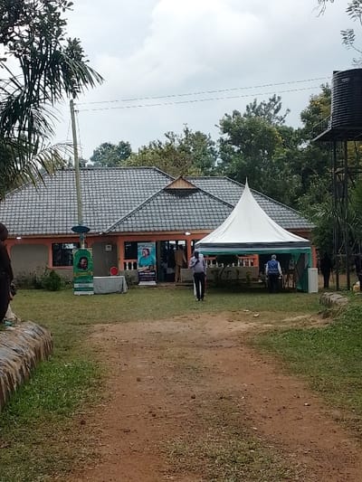 The scene depicts an outdoor gathering in the Marenyo-Shianda ward, Kenya. A single-story building with peach-colored walls and a dark grey tiled hip roof stands centrally, featuring decorative wooden accents. Its entrance is open, flanked by windows and two vertical banners. The left banner is green, displaying a man's portrait. The right banner is light blue, featuring a woman's portrait and text including "MUGWA ANNA KEDAM."
A white, pointed canopy tent with green and dark blue striped valances is positioned on the grassy area in front of the building, to the right of a reddish-brown dirt path. Several individuals are present: one man in a light blue shirt and dark trousers walks on the dirt path towards the building, to the right of the tent. Two people are partially visible inside the tent, one wearing a light blue vest. Other individuals are visible near the building's entrance, and partially obscured on the far left and right.
A tall black water tank on a metal stand is visible on the far right. A utility pole with wires stands on the left. A small round table with a white cover and a red object on top is located to the left of the building's entrance. The ground consists of a central dirt path surrounded by green grass. Lush green trees are visible in the background and on the sides. The sky is overcast.