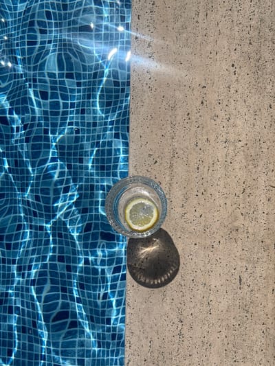 This is an overhead, top-down shot of a glass of water with a lemon slice on the edge of a swimming pool. The pool is tiled with small, square blue tiles that are shimmering with light from the sun reflecting off the water. The edge of the pool is made of a textured, light-colored stone. A dark shadow from the glass and possibly something else is cast on the stone. The water appears clear and inviting. The lighting suggests it is daytime, likely sunny, and the setting is a pool area, possibly a resort or a private residence. No people are visible in the image. There is no visible text. The location is Beşiktaş, Turkey.