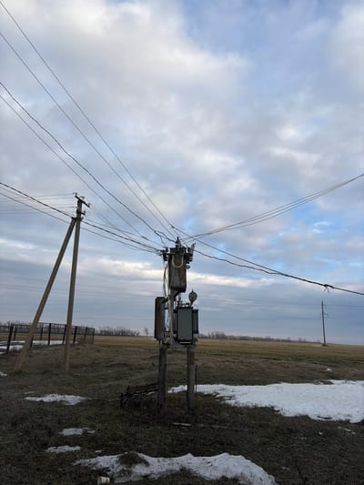 An outdoor scene depicts an electrical transformer mounted on poles, with power lines extending across a cloudy sky. A field with remnants of snow is adjacent to the infrastructure. A utility pole with angled support is visible, along with a fence line. The horizon is punctuated by distant trees.
