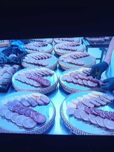 The image depicts a close-up, slightly blurry view of a buffet or food preparation area, likely indoors. Multiple circular, silver platters are filled with sliced meats and possibly other food items. The platters are arranged on a dark-colored surface. In the foreground, a person's hands, wearing black gloves, are visible handling some of the food. To the left, there's a tray containing more sliced meat. The lighting is somewhat harsh, creating reflections on the metallic platters. The overall scene suggests a catered event or a professional kitchen setting. There is no visible indication of the city of Pavlohrad, Ukraine, within the image itself.