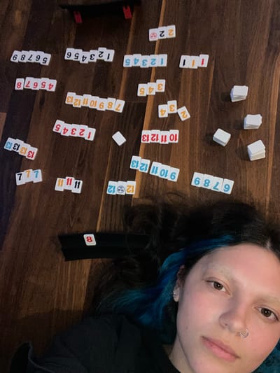 A woman with blue-tinged dark hair lies on a dark wooden floor beside Rummikub tiles arranged in runs and groups. A black tile rack with an "8" tile sits near her shoulder. The tiles include runs of 1-13 in various colors, groups of the same number, and some loose tiles. The floor's surface appears to be a dark, stained wood, and the room is dimly lit. The woman is looking at the camera with a neutral expression and has a nose ring. There is no information in the image to suggest that the location is in the city of Villaviciosa, Spain.