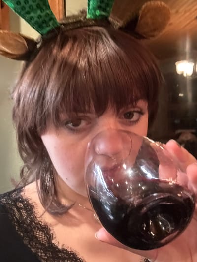 A person with dark brown hair, bangs, and reindeer antlers poses, holding a wine glass filled with dark red wine up to their nose. Their eyes are large and focused. They are wearing a black lace top. The background is dark and slightly blurry, with a possible light fixture visible. The image is at a tilted angle.