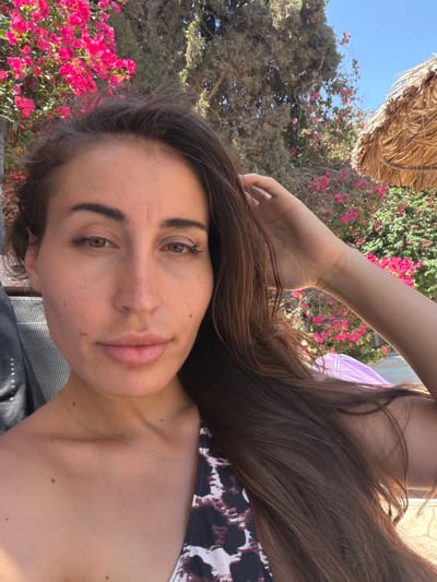 A selfie of a woman in Mazotos, Cyprus, shows her face, shoulders, and chest. She has long brown hair and is wearing a leopard-print swimsuit. She is outdoors, with bright pink flowers and trees in the background. A straw umbrella is visible in the upper right corner. The woman is touching her hair with her right hand.