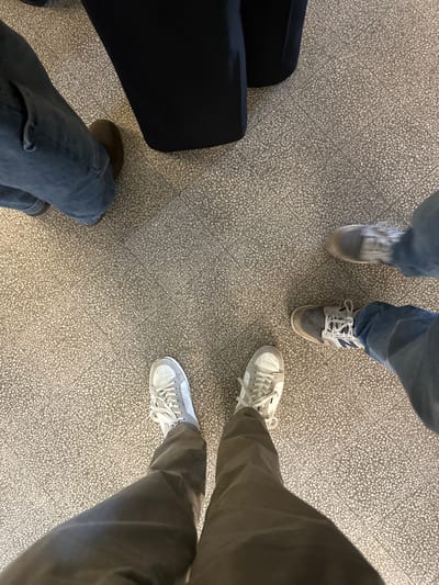 The scene depicts an overhead view of four individuals' lower legs and feet on a speckled light gray terrazzo floor with a faint grid pattern. The location is identified as the Aveiro train station in Aveiro, Portugal, based on the distinctive flooring.
From the bottom, a person is visible wearing dark brown or grey trousers and light grey and white sneakers with white laces. To the right, another person wears blue denim jeans and grey and blue athletic shoes with white laces, featuring a prominent 'N' logo. In the top-left quadrant, a third person is wearing blue denim jeans and a single dark brown shoe. The fourth individual, partially obscured in the top-right, has the hem of dark trousers or a skirt visible, but their footwear is not clearly discernible. All individuals appear to be standing on the patterned tiled floor. There are no direct interactions or specific actions visible beyond their static stances.
