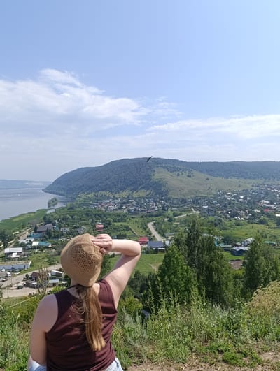 A person wearing a brown, crocheted hat and a maroon tank top is standing on a hillside, looking out over a valley. The person's right arm is raised, with their hand shielding their eyes from the sun. The valley below contains a river, a town with numerous buildings, and rolling, tree-covered hills in the distance. A bird is visible flying in the sky above the town. The sky is blue with scattered white clouds. The foreground is dominated by green vegetation and dry, rocky ground. The scene appears to be the city of Solnechnaya Polyana, Russia, likely taken from a scenic overlook.