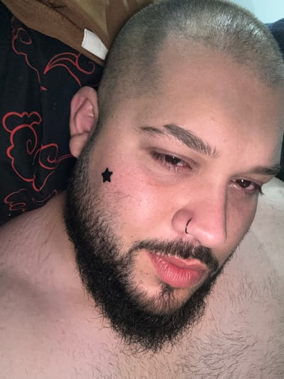 A close-up, overhead shot shows a person with a buzzed haircut and a beard resting on a pillow. The person has a black star sticker on their left cheek, a black nose ring, and their eyes are half-closed. The pillow has a black fabric with red swirling patterns. The lighting is dim, and the image is slightly blurry. The location in the city of Smyadovo, Bulgaria is not depicted in this image.