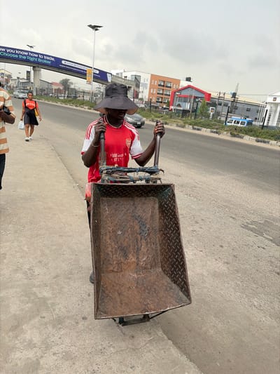The image presents an urban street scene in Jakande, Nigeria, under a light grey, overcast sky. In the foreground, a dark-skinned individual, appearing male, is positioned behind a large, empty, two-wheeled handcart. He wears a red short-sleeved jersey with white trim (an Arsenal football club shirt with "Emirates Fly Better" sponsor), dark trousers, and a dark, wide-brimmed hat that casts a shadow over his face. His hands grasp the dark metal handles of the cart, which features a rusted, dark brown diamond-plate metal bed.
To the immediate left, a portion of a second individual is visible, wearing a striped tan and white short-sleeved shirt. Further along the concrete sidewalk, a third individual, a dark-skinned female, walks away from the viewer. She wears an orange short-sleeved top, black shorts, and white sneakers, carrying a white plastic bag in her left hand.
The scene includes a multi-lane paved road running parallel to the sidewalk. A pedestrian bridge with an overhead gantry sign spans the road in the mid-ground. The sign displays prominent white text: "NOW YOU KNOW HOW TO TRADE. CHOOSE THE RIGHT BROKER." Various multi-story buildings, some residential-style with orange and white facades, and others commercial with a red roofline, are visible across the road. Vehicles, including a light-colored sedan and a white and blue van, are present on the road in the distance. Streetlights on poles are also visible above the road.