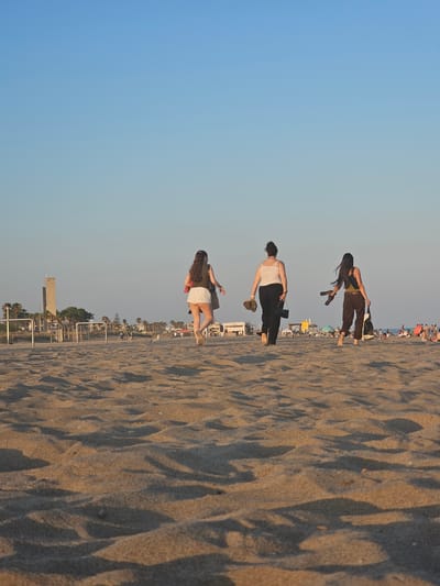 Three women walk barefoot on a sandy beach towards the water at sunset in Castelldefels, Spain. The woman on the left wears a white top and white shorts, and carries a brown purse. The woman in the middle wears a white tank top and black pants, and carries a black purse. The woman on the right wears a dark tank top and dark pants, and carries a light-colored bag and a pair of dark sandals. In the background, a tall, slender tower stands near palm trees and other buildings. The sky is clear and blue, with a gradient of lighter blue towards the horizon. The sand is rippled, with shadows cast by the setting sun.