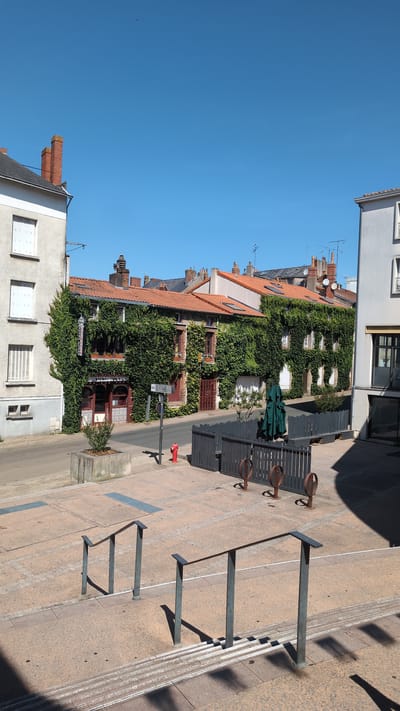 The image depicts a street scene in the city of Cholet, France, likely in a commercial or residential area with older architecture. In the foreground, a set of concrete stairs with metal handrails leads up from a tiled plaza. To the left and right of the plaza are buildings. The building on the left is a light-colored, multi-story structure with white-shuttered windows. The building in the center, which dominates the middle ground, is an older, brick building extensively covered in green ivy. It features multiple windows and an arched doorway at ground level, with signage that appears to indicate a business. A red fire hydrant is visible near the sidewalk in front of this ivy-covered building. Beyond the ivy-covered building, other buildings with red-tiled roofs and chimneys are visible. A dark grey fence with decorative circular metal elements encloses an outdoor seating area in front of one of the buildings, and a green umbrella is set up within this area. The sky is clear and blue, suggesting a sunny day. The overall impression is one of a charming, European street with historical buildings.