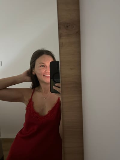 A woman is taking a selfie in a mirror. She is wearing a red slip with lace trim. She has her hair pulled back and is smiling. Her phone is visible in the mirror, and she is holding it to take the picture. The mirror is framed by a wooden border. The background is a plain white wall. The scene appears to be indoors, possibly in a bedroom or bathroom. The location in Donji Pobori, Montenegro is not discernible from the image.
