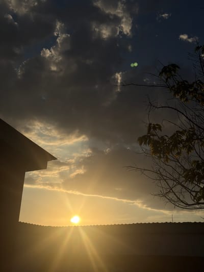 The image depicts a dramatic sky scene captured from a low angle, likely at dusk given the warm, golden light. The sun, a bright orb with visible rays, is positioned in the upper right quadrant, casting a warm glow across the sky. This light illuminates a strip of wispy clouds that stretch horizontally, contrasting with darker, more ominous clouds on the left side of the frame.
In the foreground, silhouetted against the bright sky, are the sharp angles of a building's roofline, suggesting an urban or suburban setting. To the left, branches of a leafless tree with sparse foliage hang into the frame from the top, adding a touch of nature to the scene.
A peculiar detail in the upper left quadrant is a pair of small, glowing green circles, resembling abstract shapes or perhaps a lens flare artifact. The overall mood is one of natural grandeur and perhaps a hint of foreboding due to the contrasting cloud formations.
There is no visible text in the image. The location context of Baurzhan Momyshuly, Kazakhstan, implies the scene is set in that region.