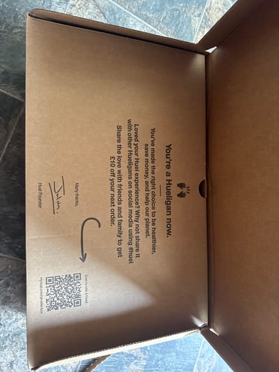 A slightly elevated eye-level shot shows the inside of an open cardboard box. The box has a brown cardboard texture with printed text and graphics. At the top center of the interior is a pair of hands clapping logo above the phrase "You're a Hueligan now". Below, it reads "You've made the right choice to be healthier, save money, and help our planet." More text follows: "Loved your Huel experience? Why not share it with other Hueligans on social media using #huel. Share the love with friends and family to get £10 off your next order." In the bottom left is the text "Many thanks," followed by a signature, "Julian." Underneath the signature is "Huel Founder". An arrow points to a QR code with "Scan to refer a friend" above it and "my.huel.com/uk-refer-box" below. The image is likely taken indoors. Huel is a nutritional food company. The domain "my.huel.com/uk-refer-box" suggests that this is a Huel box located in the United Kingdom. Desborough is not mentioned in the image and is not deducible from any of the objects or text.