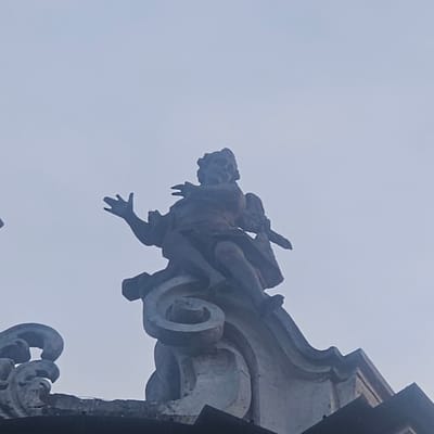 The image shows a stone statue of a winged figure, possibly an angel or cherub, seated on a decorative architectural element. The figure is depicted with its left arm raised and its right arm bent, holding what appears to be a scroll or book. The statue has a serene, yet somewhat stern expression. The architectural element it rests on features swirling, decorative patterns. The statue is silhouetted against a pale, overcast sky, suggesting it is an exterior architectural detail. The overall impression is one of aged stone, possibly weathered by time. Based on provided information, this statue is located in the city of Calcinate, Italy.