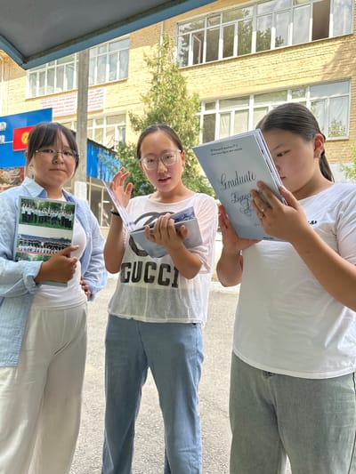 Three young women stand outside a school building in Taygaraev, Kyrgyzstan. The building has a yellow brick facade and several windows. A Kyrgyz flag is visible on the left side of the image. The woman on the left is wearing a light blue open button-down shirt over a white top and white pants. She is holding a booklet with photographs of students. The woman in the middle is wearing a white t-shirt with a black graphic and light blue pants. She is holding a booklet open and gesturing with her right hand. The woman on the right is wearing a white t-shirt and grey pants. She is holding a booklet titled "GRADUATE" and appears to be reading it. The ground is paved with asphalt.