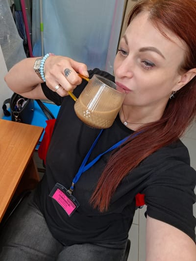 A medium shot shows a woman with reddish-brown hair drinking from a glass mug of coffee. She is wearing a black t-shirt, dark jeans, a silver watch on her left wrist with a light blue beaded bracelet, a silver ring on her left ring finger, a small pendant necklace, and a silver earring in her right ear. A pink ID badge is clipped to her t-shirt with a blue lanyard. The woman is sitting at a wooden table next to a blue surface. In the background, a mop and broom are visible. The lighting is bright and appears to be artificial. The scene is likely indoors, possibly in an office or a workplace.