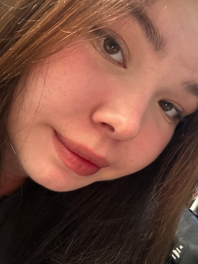 The image is a close-up, waist-up shot of a young woman with brown hair and brown eyes. She is looking towards the camera with a slight smile. The lighting suggests she is indoors, possibly in a well-lit room. The background is dark and indistinct, with some hints of her dark clothing. There are no specific landmarks or environmental cues visible to indicate her location within Surgut, Russia.