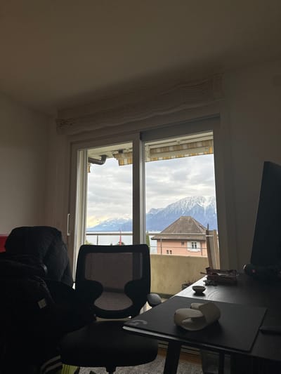 This media file captures an indoor scene looking out through a large rectangular window, offering a view of a mountainous landscape and nearby buildings.
On the interior, the window frame is prominent. To the left of the window, a white wall is visible, beneath which a textured, light-colored window covering, possibly a rolled-up blind or valance, is affixed. To the right, a dark desk is positioned, holding a computer monitor (partially seen at the top right), two small black speakers, and a dark mousepad featuring a white "G" logo. Two white, irregular-shaped decorative objects also sit on the desk. A black office chair with a mesh back is partly visible in the lower right, positioned in front of the desk. A dark jacket is draped over an unseen object below the chair.
Through the window, a picturesque exterior is revealed. Dominating the background are majestic, snow-capped mountains beneath an overcast sky. In the middle ground, a building with a reddish-brown tiled roof and light pink walls stands out. Below the mountains and buildings, a body of water, likely Lake Geneva given the location context, stretches across the horizon. A small red flag or marker can be seen near the water's edge. Closer to the window, parts of other structures are visible, including a striped yellow and white awning and a dark pipe.
The scene depicts a daytime setting with an overcast sky, providing soft, diffused light. The snow on the mountains suggests a cool or cold climate, consistent with the Swiss Alps. The visible text is the "G" logo on the mousepad, indicative of the Logitech G brand. The overall setting points to a home or office room in Corseaux, Switzerland, overlooking the alpine landscape and Lake Geneva.