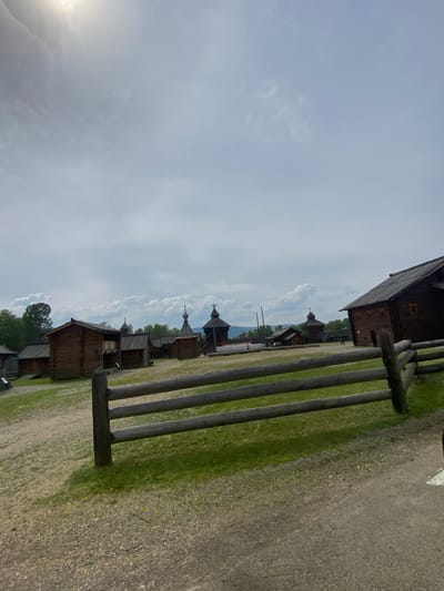The image depicts an outdoor scene in the Taltsy (Irkutsk) museum-reserve in Russia, likely during a cloudy day. In the foreground, a weathered wooden fence with three horizontal logs stretches across the frame. Beyond the fence, a dirt and gravel path gives way to patches of green grass. Several traditional wooden structures, characteristic of Siberian architecture, are visible in the mid-ground and background. These include log houses with dark wooden walls and pitched roofs, some featuring visible windows and balconies. Distinctive domed structures with spires, possibly churches or watchtowers, punctuate the landscape. In the distance, a line of trees and rolling hills can be seen under a bright, overcast sky with some cumulus clouds. A few indistinct figures, presumably visitors, are scattered throughout the site.