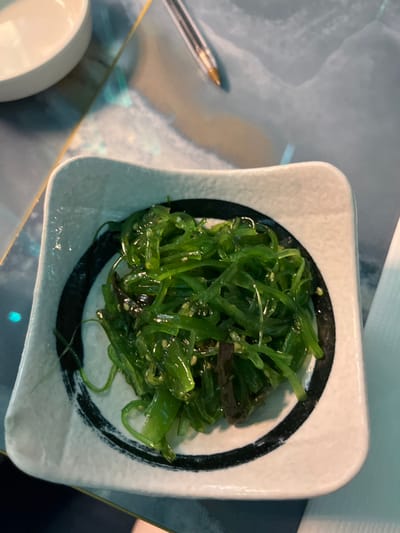 The image displays a close-up, overhead view of a small, square, white ceramic bowl filled with green seaweed salad. The bowl has a distinct black, brushstroke-like circular pattern around its inner rim. The seaweed is glossy, suggesting a dressing, and appears to be mixed with some small, light-colored particles, likely sesame seeds. A few darker, perhaps brown, pieces are also visible within the seaweed.

The bowl rests on a smooth, light-colored surface, possibly a table or placemat, which reflects light and colors. To the left of the bowl, a transparent ballpoint pen with a gold tip is visible, lying on the surface. In the bottom left corner, part of another white, rounded object, possibly a small dish or cup, is cropped out of view. The background appears to be a muted blue or gray tone, with some subtle variations in color and light, possibly indicating a reflective surface or a patterned table. The lighting suggests an indoor setting, and no specific time of day is clearly indicated. There is no visible text in the image. The food style suggests an Asian cuisine, possibly served in a restaurant or home setting in Basiglio, Italy.