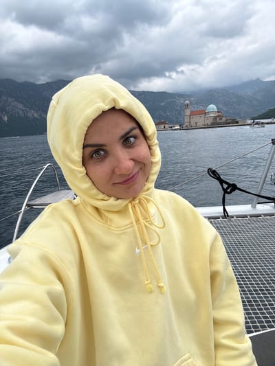 The image captures a selfie of a person on a boat, with a scenic Montenegrin landscape in the background. The photo is taken from a slightly low angle, looking up at the subject and the distant scenery.
In the foreground, a person with fair skin, dark eyebrows, and greenish-hazel eyes looks directly at the camera with a slight, content smile. They are wearing a pale yellow hooded sweatshirt (hoodie) with the hood pulled up, framing their face. White drawstrings with knotted ends and a small white Nike swoosh logo are visible on the hoodie.
The setting appears to be a boat, possibly a catamaran, given the visible white netting on the deck to the right and a metal railing. A black rope is neatly tied to the railing.
The background reveals a large body of water, likely the Bay of Kotor, with ripples on its surface. Across the water, a dramatic mountainous landscape rises steeply, characterized by dark, rocky slopes. Nestled against the mountains, or on a small island, is a distinctive building with a light blue dome and a reddish-brown tiled roof, along with a separate tower-like structure. This architectural style and placement strongly indicate the area around Perast, Montenegro, likely featuring Our Lady of the Rocks island.
The sky is overcast with thick, grey clouds, suggesting a cloudy day, but daylight is evident. The overall mood conveyed by the person's expression and the serene waters seems calm and pleasant, despite the overcast conditions.