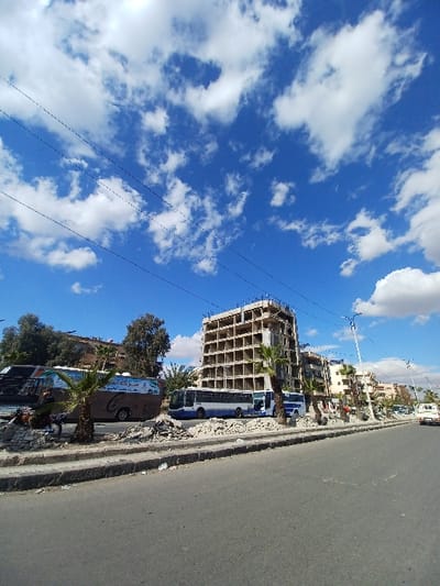 The image captures an urban street scene within Al-Qadam Municipality, Syria, under a bright blue sky dotted with scattered cumulus clouds. Utility lines are visible traversing the upper left quadrant of the sky. In the foreground, an asphalt road extends towards the mid-right, bordered by a concrete curb. Adjacent to the curb is a raised median or sidewalk area extensively covered with piles of construction rubble, fragmented concrete, and soil.

A prominent feature in the midground is a multi-story building, approximately seven to eight stories tall, in a state of construction. Its concrete skeleton, revealing floor slabs and column grids, is visible, with numerous open rectangular apertures for windows or doorways. Temporary scaffolding or support structures are discernible on its exterior.

To the left of the construction site, a large passenger bus, primarily tan and blue with Arabic script on its side, is parked on the debris-strewn median. A dark-clothed individual is observed near the front left side of this bus, appearing to be kneeling or working on the ground amidst the rubble. Several small palm trees are interspersed along the median. To the right of the tan bus and closer to the construction, another large passenger bus, white and blue, is parked. Further down the street, beyond the primary construction, a sequence of multi-story buildings, varying in architectural style and condition, lines the road. A white pickup truck is visible on the road in the far right background, facing away from the viewer. The scene is illuminated by direct sunlight, casting shadows, indicating clear daytime conditions.
