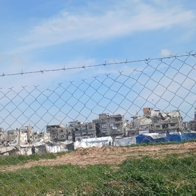 The image depicts a scene in Jabalia, Palestinian Territory, characterized by extensive structural damage and temporary habitation. In the foreground, a chain-link fence with a single strand of barbed wire running horizontally across its top edge spans the width of the frame. Below the fence, green vegetation, likely grass, transitions into an area of reddish-brown, disturbed earth. The mid-ground features several temporary shelters, including at least six white rectangular tents and two blue tents, erected on this ground amidst scattered debris. One dark-clad individual is observed walking from left to right in the lower-left mid-ground, near a white tent and a pile of light-colored rubble. In the background, a dense cluster of multi-story concrete buildings stands in various states of severe destruction, exhibiting missing walls, exposed rebar, and collapsed sections. The sky above is light blue with scattered white clouds.
