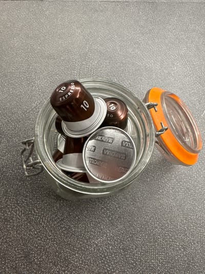 A high-angle, close-up shot captures a glass jar with an orange-tinted lid, containing several brown and silver coffee capsules. The jar is placed on a textured, grey surface. One of the brown capsules prominently displays "10 ESPRESSO" in white lettering. Another capsule, on top, shows "10" and the word "SO". A silver capsule at the bottom of the jar has a circular foil top with the word "MARCILLA" printed in an arc. The lid of the jar is slightly ajar, revealing the capsules inside. The scene is devoid of any human presence or activity.