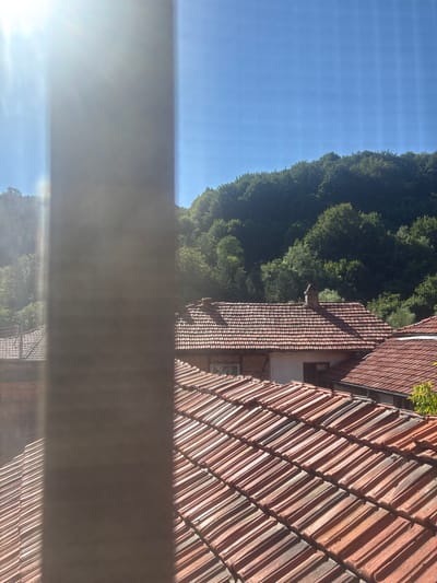 A bright sun shines from the upper left corner, casting lens flare across the image. A vertical, blurry pole or pillar obscures the center of the frame. Beyond the pillar, the foreground is dominated by an expanse of reddish-brown tiled roofs, characteristic of traditional Bulgarian architecture. In the middle ground, more tiled roofs are visible, some with brick chimneys. Behind the houses, a dense, green forest covers a sloping hill, leading up to a clear blue sky. The image appears to be taken from an elevated vantage point, possibly a window or balcony, looking out over a village in Shipkovo, Bulgaria.