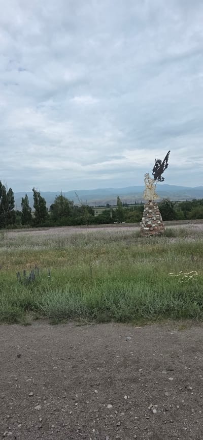 This image depicts an outdoor scene with a metal sculpture of a bird on a stone pedestal in the foreground. The sculpture is positioned in a field of wildflowers, with tall green grass in the immediate foreground. In the background, there are trees, rolling hills, and a cloudy sky. The scene appears to be located in a rural area, possibly near Sasyk-Bulak, Kyrgyzstan, given the description. There are no people visible in the image. The overall atmosphere is serene and natural.