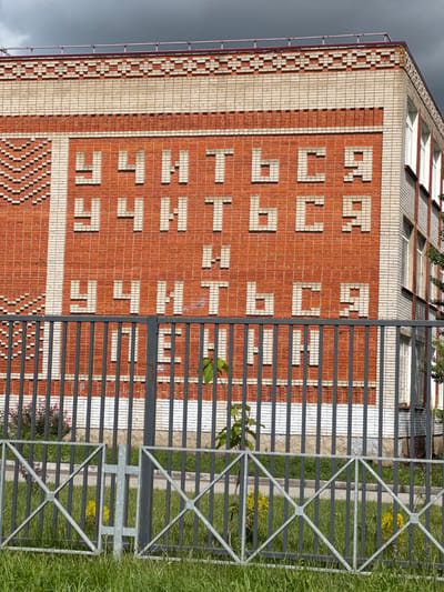 A building facade in Yelizavetinskaya, Russia, features a large inscription in Cyrillic script formed by contrasting light-colored bricks within a red brick wall. The inscription reads "ДЕТСКИЙ САД," which translates to "Kindergarten". To the right of the building, a metal fence encloses a grassy area. The sky is overcast. The building is constructed from bricks of different colors. There is greenery growing next to the fence.