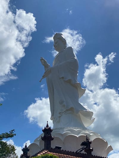 The image shows a low-angle, full-body shot of a large, white statue of a woman from the waist up, set against a vibrant blue sky dotted with fluffy white clouds. The statue is positioned on the right side of the frame, with its head and upper torso tilted towards the left. The woman is wearing a flowing, draped garment that gives the impression of movement, with its folds and curves highlighted by the sunlight. Her left arm is extended outwards and downwards, with her hand open and fingers spread, while her right arm is bent and held closer to her body. The statue appears to be made of a smooth, white material, possibly stone or plaster, and shows intricate details in the folds of the clothing.
Part of a darker, ornamental structure is visible to the right of the statue, possibly part of its base or a surrounding building. This structure features decorative elements, including pointed spires and dark, wrought-iron-like fixtures. In the upper right corner of the image, some greenery is visible, suggesting trees or foliage.
The sky is a clear, bright blue, indicating it is daytime. The presence of scattered cumulus clouds suggests fair weather, possibly with a gentle breeze. The perspective from below the statue, looking upwards, emphasizes its immense size and grandeur. The overall mood is one of serenity and perhaps reverence, evoked by the peaceful posture of the statue and the vastness of the sky. The image appears to be taken outdoors, likely in a public space or religious site in Thôn 5, Vietnam, given the provided location context. No text is visible in the image.