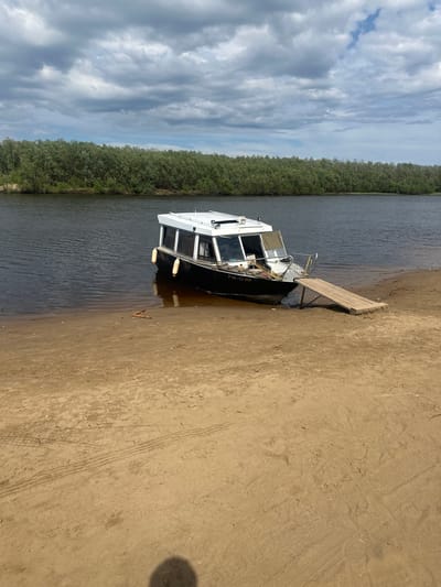 A riverboat is partially beached on a sandy shore, with a wooden ramp extending from the boat to the land. The boat is dark-hulled with a white cabin and appears to be moored. Two yellow fenders hang from the boat's side. In the background, a dense line of green trees borders the dark water of the river. The sky above is filled with dramatic, dark gray clouds, with patches of blue sky visible. The sandy shore shows tire tracks and some debris. The scene is calm and appears to be in a natural, undeveloped area. The license plate on the boat reads "P 18-52 III".