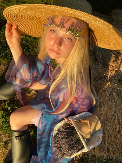 A young person with long blonde hair lies on their back, tilted to the right. They are wearing a large, straw sun hat and a flowing, sheer iridescent dress in shades of blue and purple. Delicate sprigs of lavender and small white flowers adorn their hair near their temple. Their eyes are open and looking directly at the camera. Beside them is a small white wicker basket filled with dried lavender. The person is lying on a bed of hay, with grass visible beneath. The scene is bathed in warm, golden sunlight, suggesting it is late afternoon or early evening. The setting appears to be outdoors, in a rural or natural environment, possibly a field. The overall mood is serene and ethereal.