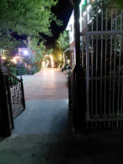 The image depicts a dimly lit outdoor scene, likely at night, with decorative lighting. A metal gate with intricate patterns forms the foreground on the left side, partially obscuring a path leading into the distance. The path appears to be paved with a textured material and is illuminated by warm, yellowish lights, possibly string lights or lanterns, that are draped around trees and structures. Lush green foliage, some adorned with lights, flanks the path, creating a festive and somewhat magical atmosphere. In the background, hints of buildings or structures can be seen, also illuminated by various lights. The overall impression is one of a beautifully decorated outdoor space, possibly a garden or a public area, set up for an evening event or celebration. The time of day is clearly night due to the darkness and the reliance on artificial lighting. The location context suggests this is in Ақши, Kazakhstan. There are no people visible in the frame, and no text is discernible.