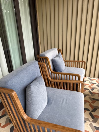 Two light blue cushioned armchairs with natural wooden frames and slatted details are positioned on a geometrically tiled patio in Púng Luông, Vietnam. The chair in the foreground, partially visible on the left, features a rectangular throw pillow. Behind it, a second, more completely visible chair mirrors its design. To the left, a dark-framed glass door or window with sheer white curtains reflects ambient light. To the right, a textured wall with vertical panels in a beige hue extends upwards. The floor features a multi-colored geometric tile pattern in shades of cream, terracotta, dark gray, and light gray. The scene is illuminated by natural daylight. No individuals are present.