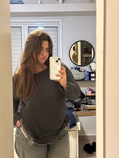 A woman in a gray long-sleeved shirt and jeans takes a selfie in front of a mirror. Her brown hair falls across her face. The mirror reflects a room with a round mirror on the wall, a window with blinds, and cluttered shelves. Black shoes hang on a closet rod in the background. The woman is holding a white iPhone in her right hand.