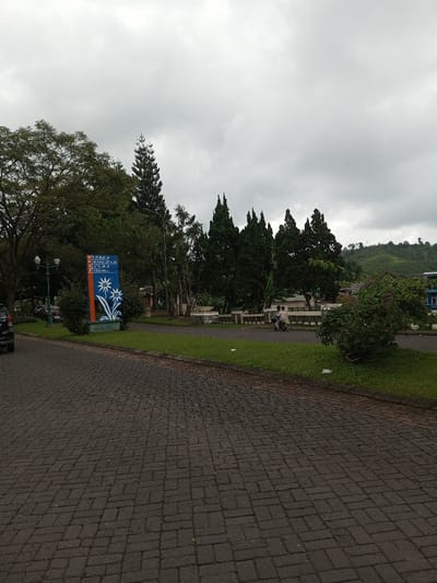 A landscape features a brick road leading to a park area with trees, grass, and a low concrete fence. A sign stands to the left with blue and orange colors and white flower images, possibly a park directory. People on a scooter are visible further back. The sky is overcast with dull grey clouds. A small hill is visible in the distance on the right.