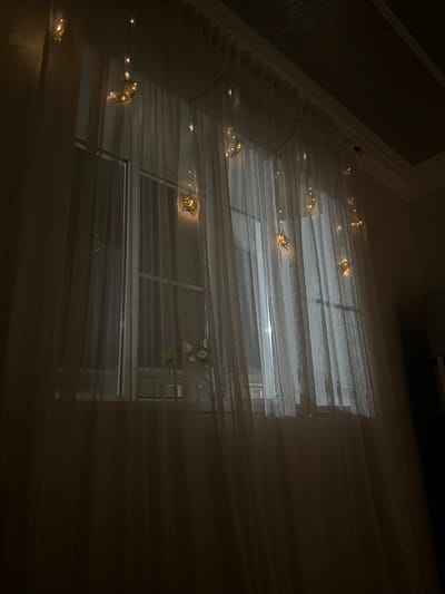 The image shows a window covered by sheer, white curtains. A string of decorative lights with crescent moon and star shapes is draped across the top of the curtains, illuminating them with a warm glow. The lights are a prominent feature against the dark interior and the dimly lit window. A vase with a few flowers is visible on a surface behind the curtains, suggesting the window is in a room. The overall ambiance is calm and dimly lit, with the light from the string decorations creating a gentle illumination. The location is described as Paraul, Russia.