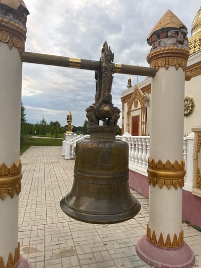 The image depicts a large, antique brass bell with intricate carvings, prominently displayed in what appears to be a temple or spiritual complex. The bell is mounted on a metal stand, with ornate golden decorative elements adorning the surrounding white columns. One of these columns shows signs of damage, with its decorative facade crumbling and revealing brickwork beneath.

In the background, a paved walkway extends towards a traditional-looking building with a red door and a white balustrade. Beyond the building, a lush green landscape with trees and a small, golden spire can be seen under a cloudy sky, suggesting an overcast day. The scene seems peaceful and serene, with no people visible. The overall impression is one of historical and cultural significance, perhaps a place of worship or a museum dedicated to religious artifacts. The location is identified as Petrovo, Russia.