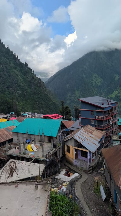 This is a vertical, eye-level, outdoor, full shot taken in Malana, India. The image captures a dense settlement nestled within a valley, with steep, tree-covered mountains flanking either side. The sky is overcast with dramatic clouds interspersed with patches of blue.
The foreground is dominated by the rooftops of various buildings, displaying a mix of construction materials and architectural styles. Several structures have bright, corrugated metal roofs in teal and red. Others are in various stages of construction, with exposed concrete pillars and reinforcement bars. One building, a multi-story structure with a dark blue facade and red balconies, stands out against the more rustic dwellings. A narrow, winding pathway snakes through the settlement, with some residents visible.
In the midground, more houses are visible, some with rusted metal roofs and others with colorful painted walls. There are piles of materials and some agricultural activity, including a patch of green corn stalks in the lower right corner.
The background features the imposing, densely forested slopes of the Himalayan mountains, disappearing into the clouds. The overall impression is of a remote, high-altitude village life.
