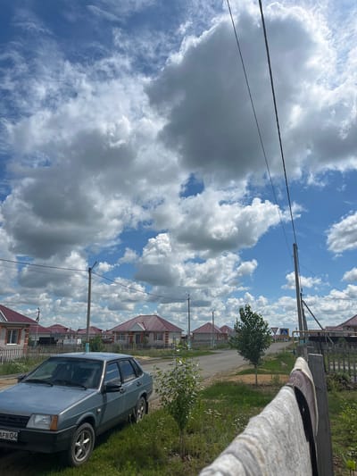 The image depicts a residential street in the village of Qobda, Kazakhstan. In the foreground, a light blue sedan is parked on the grassy shoulder of a paved road. The car is facing left, with its front visible. To the right of the car, a wooden fence runs along the property line, with a light-colored blanket or towel draped over its top. Beyond the fence, a small tree with green leaves stands next to the road. The road stretches into the distance, lined with houses that have red-tiled roofs and light-colored walls. A few streetlights are visible along the street. The sky is a vibrant blue, filled with large, fluffy white clouds. Several power lines are strung across the scene, supported by utility poles. In the background, road signs are visible, including a pedestrian crossing sign and a yield sign. The overall scene suggests a quiet, suburban or rural setting on a partly cloudy day.