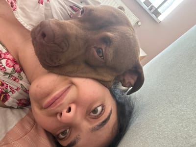 A close-up, low-angle selfie shows a person lying on a bed with a brown dog resting its head on the person's upper chest and neck. The person is looking at the camera with their eyes open and mouth slightly ajar. The dog's head is positioned on the person's shoulder, with its nose touching the person's neck and its eyes looking slightly towards the camera. The person is wearing a floral patterned top and is lying on a light-colored bedspread. The background shows a portion of a wall, a window with blinds, and a light-colored upholstered surface, possibly a couch. The scene is indoors, in what appears to be a domestic setting. There is no indication of the city of Malevo, Bulgaria, or any specific urban context.
