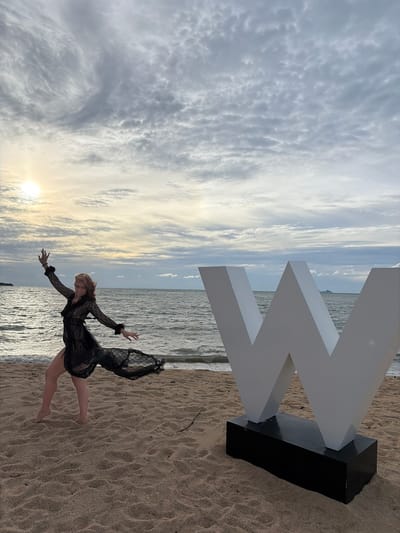 A woman in a black lace dress dances on a sandy beach in Ko Samui District, Thailand. The sun is setting, casting a warm glow on the scene. A large, white "W" sculpture stands to the right of the woman. The ocean stretches out behind them, with a cloudy sky overhead.