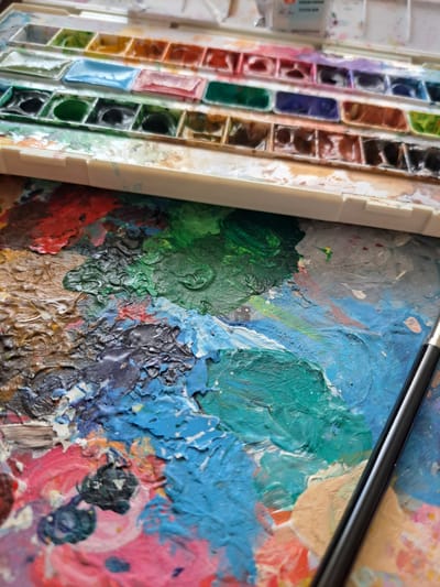 This image captures a close-up view of an artist's palette and a watercolor paint set. The subjects are the art supplies themselves, arranged on a surface that appears to be a wooden palette covered in thick, textured paint. The paint set on the left is filled with numerous rectangular pans of watercolors in various colors, including reds, oranges, yellows, greens, blues, purples, and browns. Some of the pans show signs of use, with pigment worn down, while others appear less touched.

The palette on the right is a riot of color and texture. Thick globs of paint, predominantly blues, greens, pinks, and browns, are mixed and layered, creating a tactile, almost sculptural surface. The paint application is uneven, with peaks and valleys formed by the brushstrokes. A dark, slender paintbrush rests diagonally across the palette, its tip likely loaded with pigment. The overall impression is one of an active art studio, with the tools of creation laid out and ready for use.

There are no people visible in the image. The scene is static, focusing solely on the art materials. The time of day and weather are not discernible. There are no specific location cues beyond the fact that the image was taken in Popovo, Bulgaria, which is not visually represented in the image itself. The emotions evoked are those associated with creativity and artistic process – perhaps anticipation, focus, or the satisfaction of a completed stage of work.

No visible text is present within the main composition, though there might be labels on the watercolor pans or tubes that are too small or out of focus to read. The emphasis is on the visual elements of color, texture, and the arrangement of artistic tools.